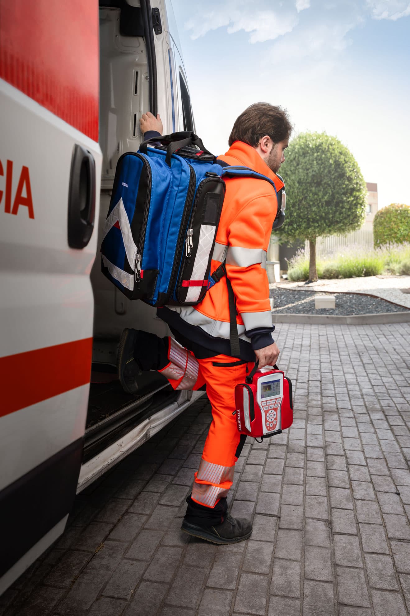 Paramedic in orange gear carrying a blue backpack and medical device into an ambulance.